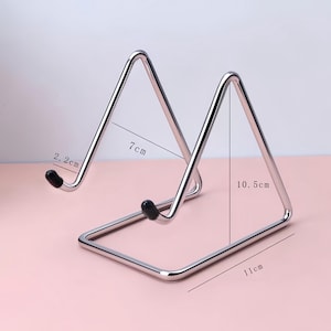 May include: A silver-colored metal display stand with a triangular frame and black rubber tips on the front hooks. The stand features printed dimensions of 2.2 cm, 7 cm, 10.5 cm, and 11 cm, indicating its physical size for holding various items.