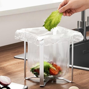 May include: A silver metal kitchen waste holder with a square frame and four legs, featuring a clear plastic bag draped over the top edges. The bag contains food scraps, and a piece of green lettuce is being placed inside the container.