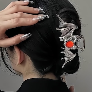Puede incluir: Una pinza para el cabello de tono plateado con forma de murciélago con las alas extendidas. El centro del murciélago presenta una piedra preciosa roja de forma ovalada y lisa. La pinza cuenta con un mecanismo de resorte para sujetar el pelo.