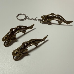 Puede incluir: Llaveros y amuletos de esqueleto de pez de color bronce. Los esqueletos de pez tienen bocas abiertas y estructuras de costillas detalladas. Un llavero tiene una cadena y un anillo. Los otros dos son amuletos. Los artículos están sobre una superficie blanca.