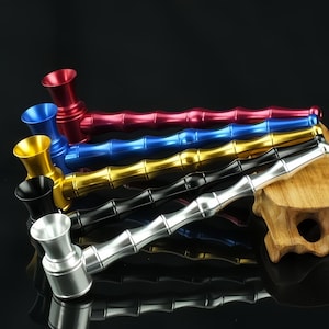May include: A collection of five metal smoking pipes arranged in a stack. Each pipe features a long, segmented stem resembling bamboo and a wide, flared bowl. The pipes are finished in metallic red, blue, gold, black, and silver colors.