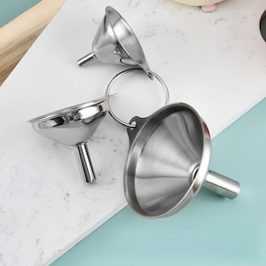 May include: A set of three silver-colored stainless steel funnels of varying sizes, each featuring a wide conical top and a narrow cylindrical spout. The three funnels are connected together by a single circular metal ring through small tabs.