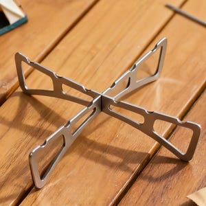 May include: A silver-colored metal cross-shaped camping stove stand featuring four legs with triangular cutouts and notched edges. The structure is composed of two interlocking pieces that form a stable base for cookware during outdoor cooking.