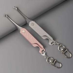 Może przedstawiać: Dwa metalowe narzędzia do czyszczenia uszu z końcówką w kształcie łyżeczki i płaskim, teksturowanym uchwytem. Jedno narzędzie jest w kolorze różowego złota, a drugie srebrne. Każdy uchwyt posiada wycięcie otwieracza do butelek i karabińczyk.