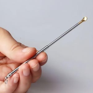 May include: A slender, telescopic metal tool featuring a polished silver-toned body and a small, rounded gold-toned scoop tip. The base of the handle includes a small circular ring attachment, allowing for easy hanging or storage of the instrument.