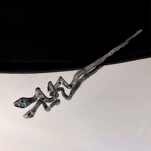 May include: A hair stick featuring a metallic, dark grey, winding snake design with a textured, branch-like body. The snake head is detailed with two small, vibrant green, circular eyes, and the overall form tapers into a long, pointed end.
