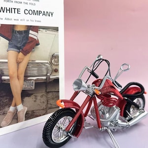 May include: A red and silver wire motorcycle model with black tires and an orange accent. The motorcycle is next to a photo with the text "WHITE COMPANY" and "-7084 CAROLINA".