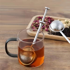 May include: A stainless steel tea infuser with a long, slender handle and a spring-loaded plunger mechanism, submerged in a clear glass mug filled with amber-coloured tea. A second identical tea infuser rests on a wooden tray with dried flowers.