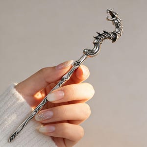 Può includere: Un bastoncino per capelli in metallo color argento caratterizzato da una testa di drago dettagliata sulla parte superiore con la bocca aperta e un corpo curvo e strutturato che si assottiglia in un'asta liscia e appuntita con motivi a onde.
