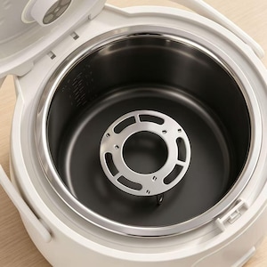 May include: A white electric rice cooker with an open lid, revealing a dark non-stick inner cooking pot. Inside the pot sits a circular, silver-toned metal steaming rack with cut-out slots, designed to hold food above the base of the cooker.