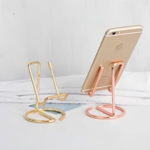 May include: Two metal wire phone stands with circular bases. One stand is gold-toned and empty, while the other is rose gold-toned and holds a gold smartphone in a vertical position. The stands feature a simple, open-frame design.