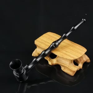 May include: A long, slender black smoking pipe with a smooth, glossy finish and a segmented design resembling bamboo. The pipe features a wide, circular bowl at one end and a tapered mouthpiece at the other, resting on a light-colored wooden stand.