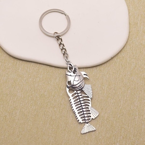 Puede incluir: Un llavero de metal plateado con un colgante en forma de esqueleto de pez. El dije está unido a una anilla circular estándar mediante una cadena corta de metal. El diseño del esqueleto de pez incluye una cabeza detallada, espina y aletas.