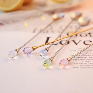 May include: A set of five long-handled metal spoons with gold-toned stems and faceted, teardrop-shaped crystal finials in various pastel colours including clear, purple, light blue, pale green, and soft pink, arranged on a white surface.