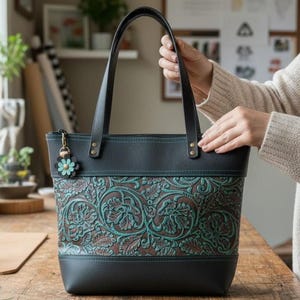May include: A black tote bag featuring a central panel with a brown and teal floral embossed pattern. The bag has black leather-like handles, a zippered top closure, and a small flower-shaped charm attached to a gold-toned metal clasp.