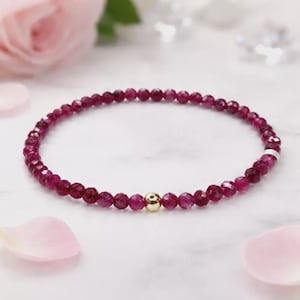 Könnte beinhalten: Ein zartes Armband mit kleinen, facettierten, tiefroten Perlen. Eine einzelne goldfarbene Perle ist in der Mitte des Armbands. Das Armband wird auf einer weißen Oberfläche mit rosa Blütenblättern im Hintergrund präsentiert.