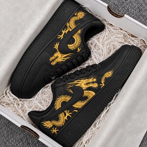 May include: A pair of black sneakers featuring a gold dragon graphic on the side panels. The shoes have black laces, a black sole, and the word AIR embossed on the heel area. The sneakers are presented inside a white box with white paper filler.