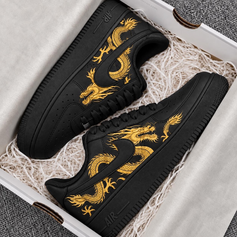 May include: A pair of black sneakers featuring a gold dragon graphic on the side panels. The shoes have black laces, a black sole, and the word AIR embossed on the heel area. The sneakers are presented inside a white box with white paper filler.