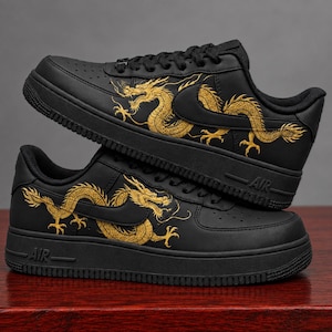 May include: A pair of black sneakers featuring a gold dragon illustration on the side panels. The shoes have black laces, a black midsole with the text AIR embossed on the side, and a perforated toe box. The dragon design wraps around the swoosh.