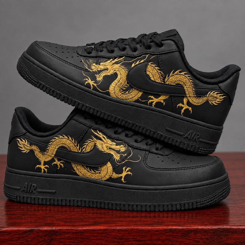 May include: A pair of black sneakers featuring a gold dragon illustration on the side panels. The shoes have black laces, a black midsole with the text AIR embossed on the side, and a perforated toe box. The dragon design wraps around the swoosh.