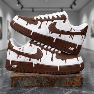 May include: A pair of white and brown sneakers featuring a chocolate drip pattern across the upper. The shoes have white laces, a brown Nike swoosh, and the word AIR embossed on the side of the brown rubber sole. The design mimics melting liquid.