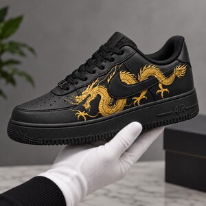 May include: A black low-top sneaker featuring a detailed gold dragon graphic along the side panel. The shoe has black laces, a perforated toe box, and a black rubber sole with the text AIR embossed on the side. The design includes a black swoosh.