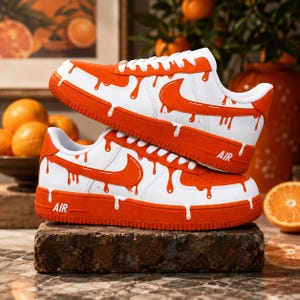 Peut inclure: Une paire de baskets blanches et orange avec un motif graphique effet goutte. Les chaussures ont des lacets blancs, des logos swoosh orange sur les côtés et le mot AIR en relief sur les semelles orange. Le design inclut des coulures orange.