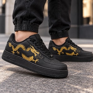 May include: A pair of black sneakers featuring a gold dragon graphic on the side panels. The shoes have black laces, a black midsole with the text AIR embossed on the side, and a black outsole. The design includes a black swoosh logo integrated into the dragon artwork.
