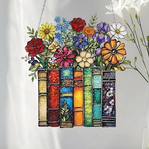 May include: A stained glass window panel featuring a colorful bouquet of flowers above a row of books. The flowers include red roses, yellow daisies, and purple irises. The books are in various colors, including red, blue, and green. The panel hangs from a silver chain.