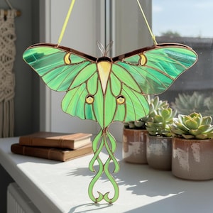 May include: A stained glass luna moth suncatcher with green wings and a brown body. The moth is hanging in front of a window. The wings have a textured appearance and are accented with yellow and brown details. The moth's body is a light yellow color.