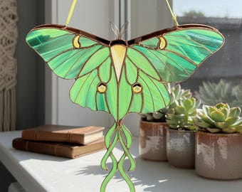 May include: A stained glass luna moth suncatcher with green wings and a brown body. The moth is hanging in front of a window. The wings have a textured appearance and are accented with yellow and brown details. The moth's body is a light yellow color.
