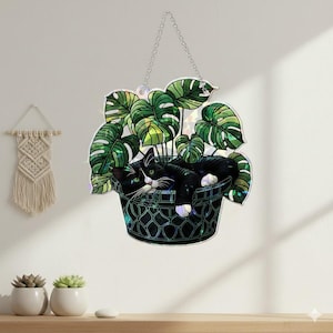 May include: A decorative hanging featuring a black and white cat resting in a green potted plant with large leaves. The cat has white paws and a white patch on its face. The plant and cat are in a dark green basket-like container, suspended by a silver chain.