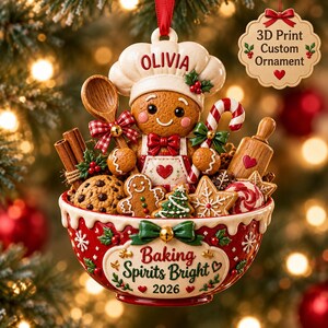 May include: A Christmas ornament featuring a gingerbread person wearing a chef hat with the name OLIVIA, inside a red bowl labeled Baking Spirits Bright 2026. The bowl contains cookies, a wooden spoon, a rolling pin, and cinnamon sticks.