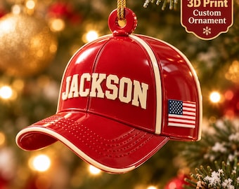 May include: A red baseball cap ornament with a cream-colored brim edge and the name JACKSON in cream lettering across the front. A small American flag patch is on the side. The ornament hangs from a gold cord and features 3D Print Custom Ornament text.