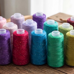 May include: A selection of colourful thread spools, each with a white top. The thread colours include pink, purple, red, blue, green, yellow, and grey. The spools are arranged on a wooden surface.