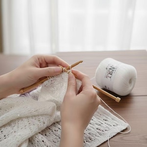 May include: A person's hands are shown knitting with wooden needles and white yarn. A partially completed white lace knit project is visible. A ball of white yarn with the text "TEN" is on the right side of the image. The scene is set on a wooden table.