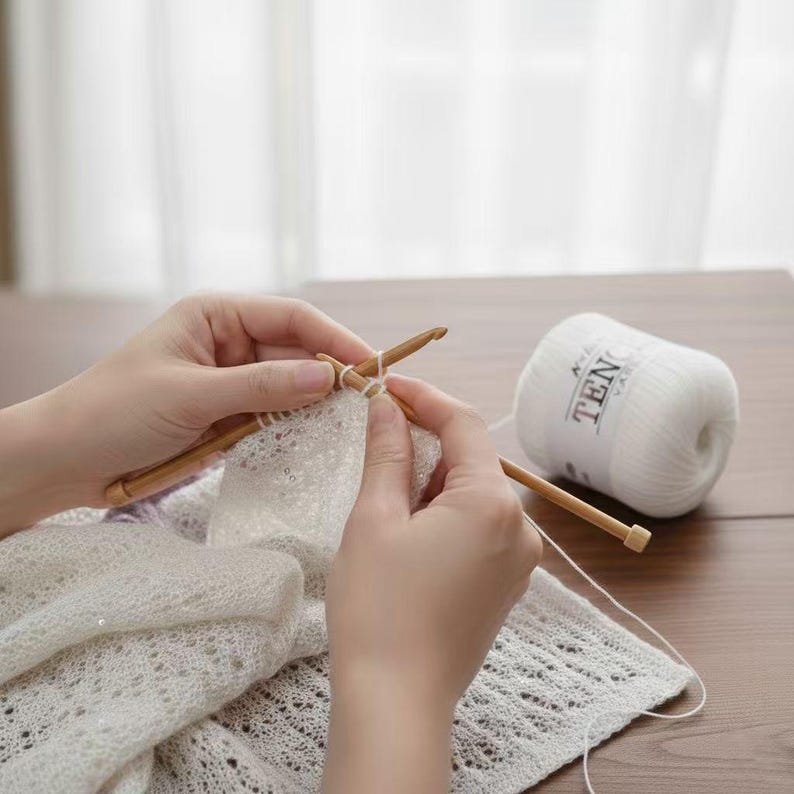May include: A person's hands are shown knitting with wooden needles and white yarn. A partially completed white lace knit project is visible. A ball of white yarn with the text "TEN" is on the right side of the image. The scene is set on a wooden table.