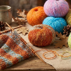 Puede incluir: Naturaleza muerta con ovillos de lana coloridos en tonos naranja, azul, rosa y verde. Se ven agujas de tejer y una pieza parcialmente tejida, junto con una taza, piñas y hojas de otoño, sugiriendo un tema otoñal acogedor.