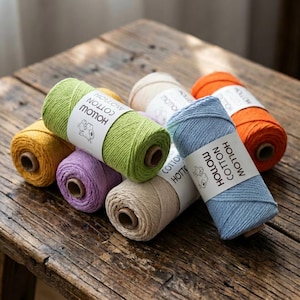 Puede incluir: Una colección de coloridas bobinas de hilo de algodón hueco en tonos amarillo, verde, morado, beige, azul y naranja. Cada bobina tiene una etiqueta con el texto "HOLLOW COTTON". El hilo se muestra sobre una superficie de madera rústica.