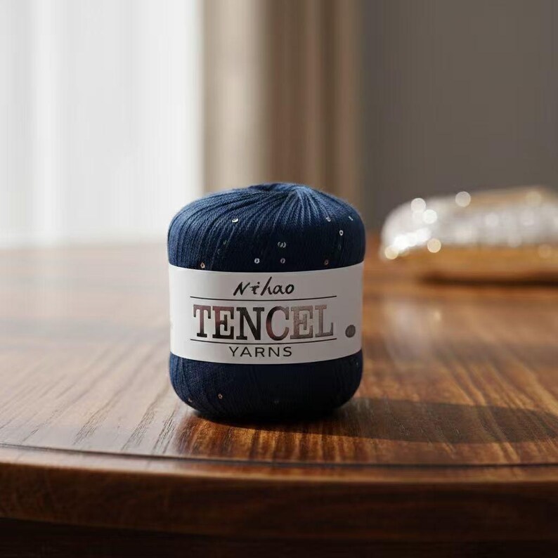 May include: A ball of navy blue Tencel yarn with a white label. The label reads "Nihao Tencel Yarns" in a bold, sans-serif font. The yarn is speckled with small, shiny embellishments, and sits on a wooden surface.