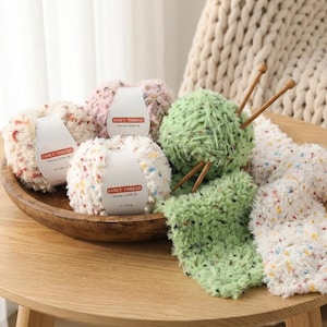 May include: A wooden bowl holds several skeins of Fancy Thread yarn in white, pink, and speckled colors. A ball of green yarn with knitting needles and a knitted swatch are also on the wooden table. The yarn is for knitting and crochet projects.