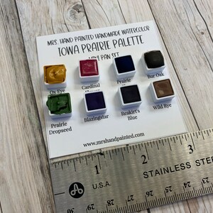 May include: A watercolor paint set with 8 colors, labeled "Ox Eye", "Cardinal", "Prairie", "Bur Oak", "Prairie Dropseed", "Blazingstar", "Reakirt's Blue", and "Wild Rye". The set is labeled "MRS. HAND PAINTED HANDMADE WATERCOLOR IOWA PRAIRIE PALETTE MINI PAN SET" and "www.mrshandpainted.com".