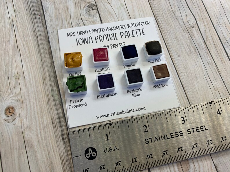 May include: A watercolor paint set with 8 colors, labeled "Ox Eye", "Cardinal", "Prairie", "Bur Oak", "Prairie Dropseed", "Blazingstar", "Reakirt's Blue", and "Wild Rye". The set is labeled "MRS. HAND PAINTED HANDMADE WATERCOLOR IOWA PRAIRIE PALETTE MINI PAN SET" and "www.mrshandpainted.com".