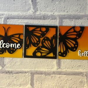 May include: A three-piece wall art set with butterfly designs. Each panel has an orange and yellow gradient background. The first panel says "welcome" in white script, and the third panel says "hello". The butterflies are black.
