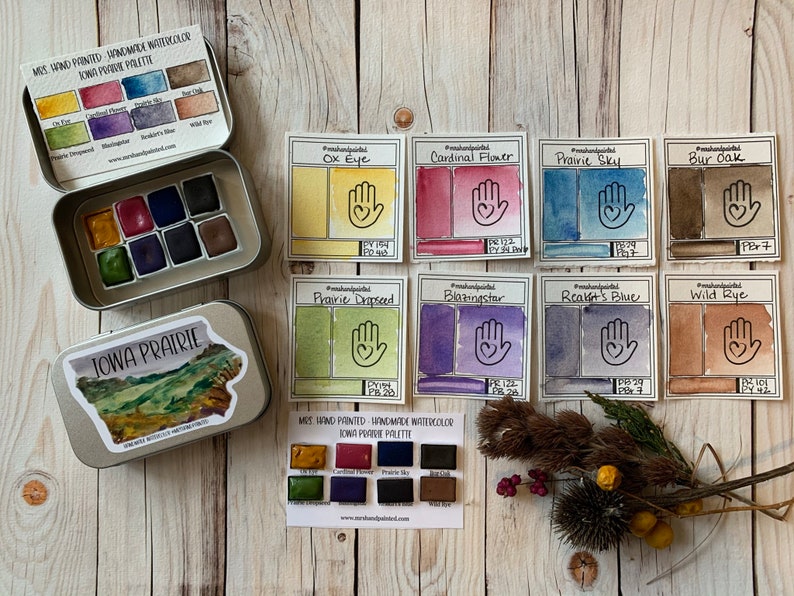 May include: A set of handmade watercolor paints in a tin box. The paints are named "Iowa Prairie" and include colors like "Ox Eye", "Cardinal Flower", "Prairie Sky", "Bur Oak", "Prairie Dropseed", "Blazingstar", "Reagan's Blue", and "Wild Rye".