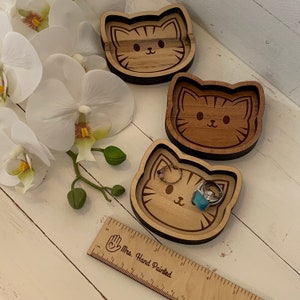 May include: Three wooden cat-shaped trinket dishes. The dishes are made of light brown wood and have a dark brown border. The cat faces are engraved on the dishes. One dish has a turquoise ring on top of it.