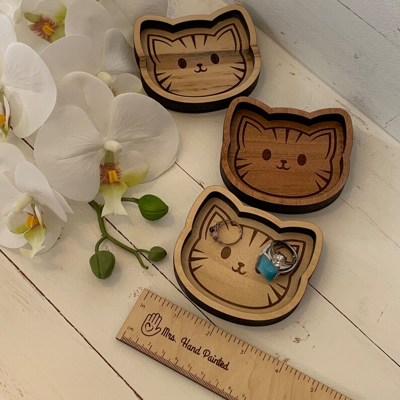 May include: Three wooden cat-shaped trinket dishes. The dishes are made of light brown wood and have a dark brown border. The cat faces are engraved on the dishes. One dish has a turquoise ring on top of it.
