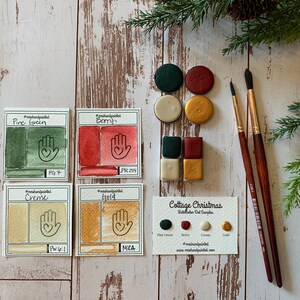 Puede incluir: Un conjunto de cuatro muestras de pintura acuarela en tonos de verde, rojo, crema y dorado. Las muestras est&aacute;n etiquetadas con sus nombres y n&uacute;meros de pigmento. El conjunto tambi&eacute;n incluye una tarjeta con los nombres de los colores y el texto "Cottage Christmas Watercolor Set Samples".