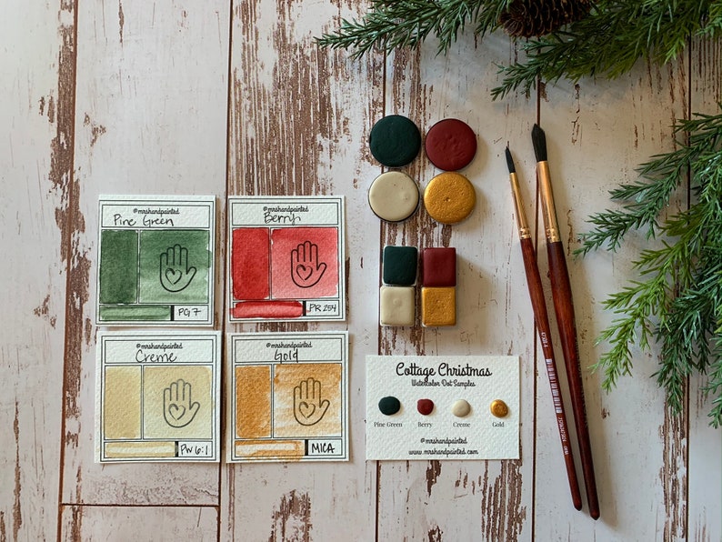 Puede incluir: Un conjunto de cuatro muestras de pintura acuarela en tonos de verde, rojo, crema y dorado. Las muestras est&aacute;n etiquetadas con sus nombres y n&uacute;meros de pigmento. El conjunto tambi&eacute;n incluye una tarjeta con los nombres de los colores y el texto "Cottage Christmas Watercolor Set Samples".