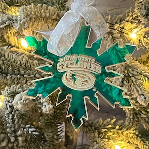 Puede incluir: Un adorno de copo de nieve verde y dorado de los Ciclones de Iowa State con una cinta blanca. El adorno cuelga de un árbol de Navidad con luces y ramas heladas. El texto "IOWA STATE CYCLONES" es visible.